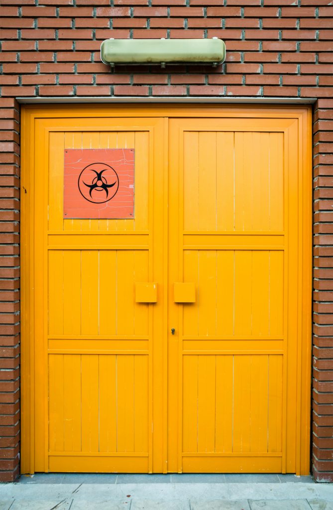 Bright yellow door with biohazard symbol sign set on a red brick wall, symbolizing caution and danger. Porte jaune vif avec un panneau de symbole de danger biologique sur un mur de briques rouges, symbolisant la prudence et le danger.