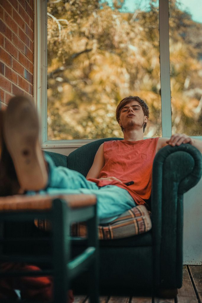 Un jeune homme en chemise orange se détend dans un fauteuil à côté d'une fenêtre ensoleillée à l'intérieur.