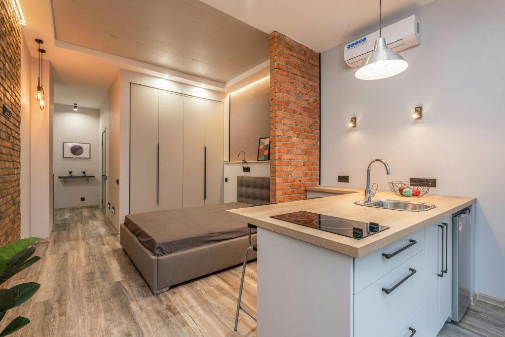 Miniature cuisine avec appareils modernes et chambre à coucher derrière une partition en brique dans un petit appartement de style loft.